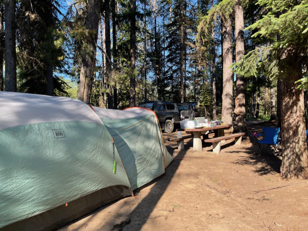 Large green and white REI tent set up at a sunny forested campsite with picnic table, truck, and blue camping chair during a high strangeness investigation in the Pacific Northwest.